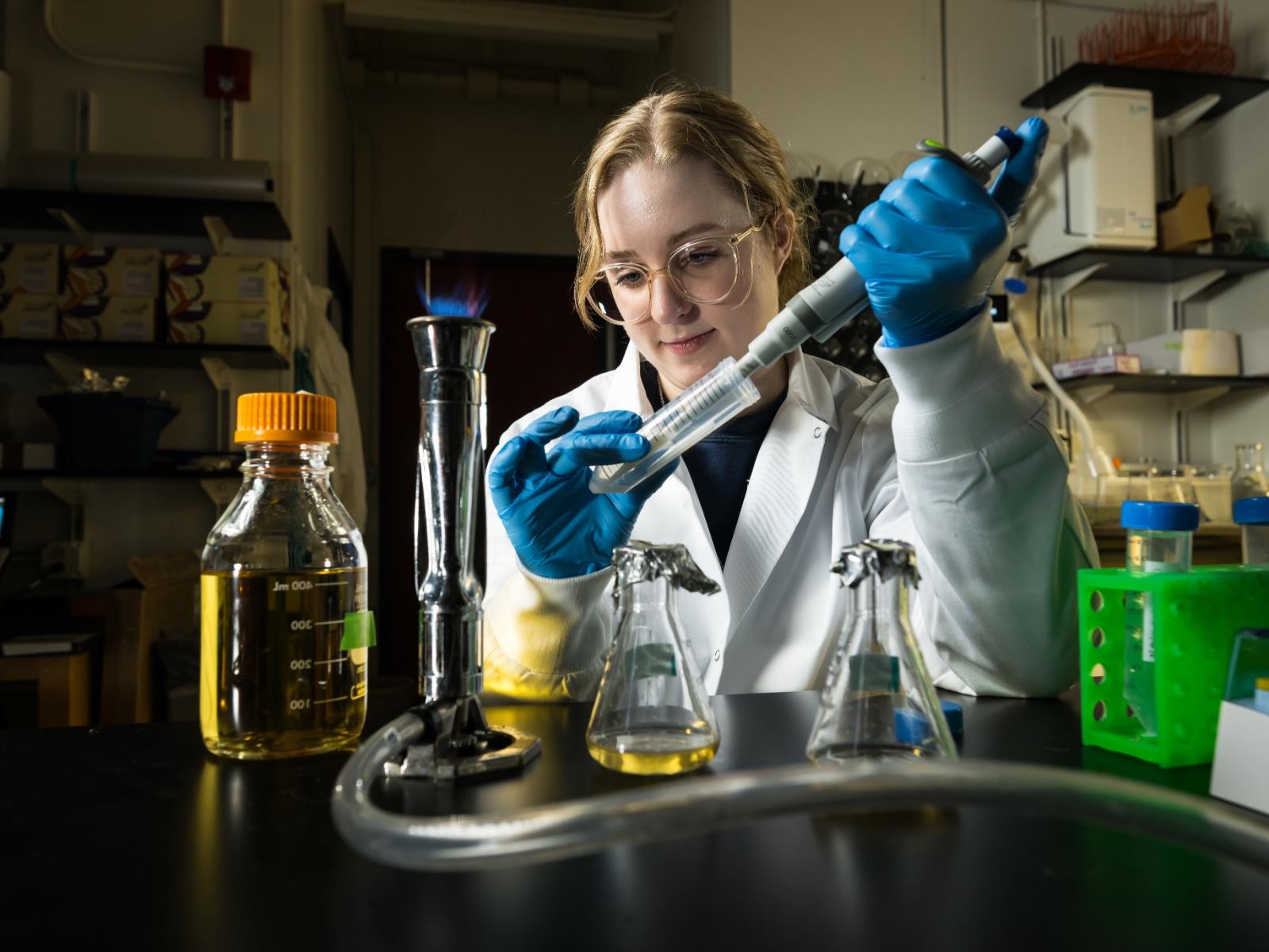 Student in labcoat sitting at desk uses equipment and test tube with various beakers in front of her